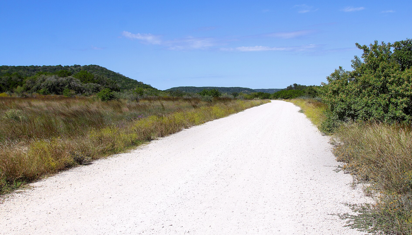 Property access and staging for a Llano County drilling quote Unpaved Hill Country access road with open roadside acreage and low wooded ridges for equipment-access review